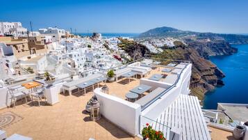 Una terraza en la azotea con mobiliario blanco, vistas a un pueblo costero y un cielo azul despejado.