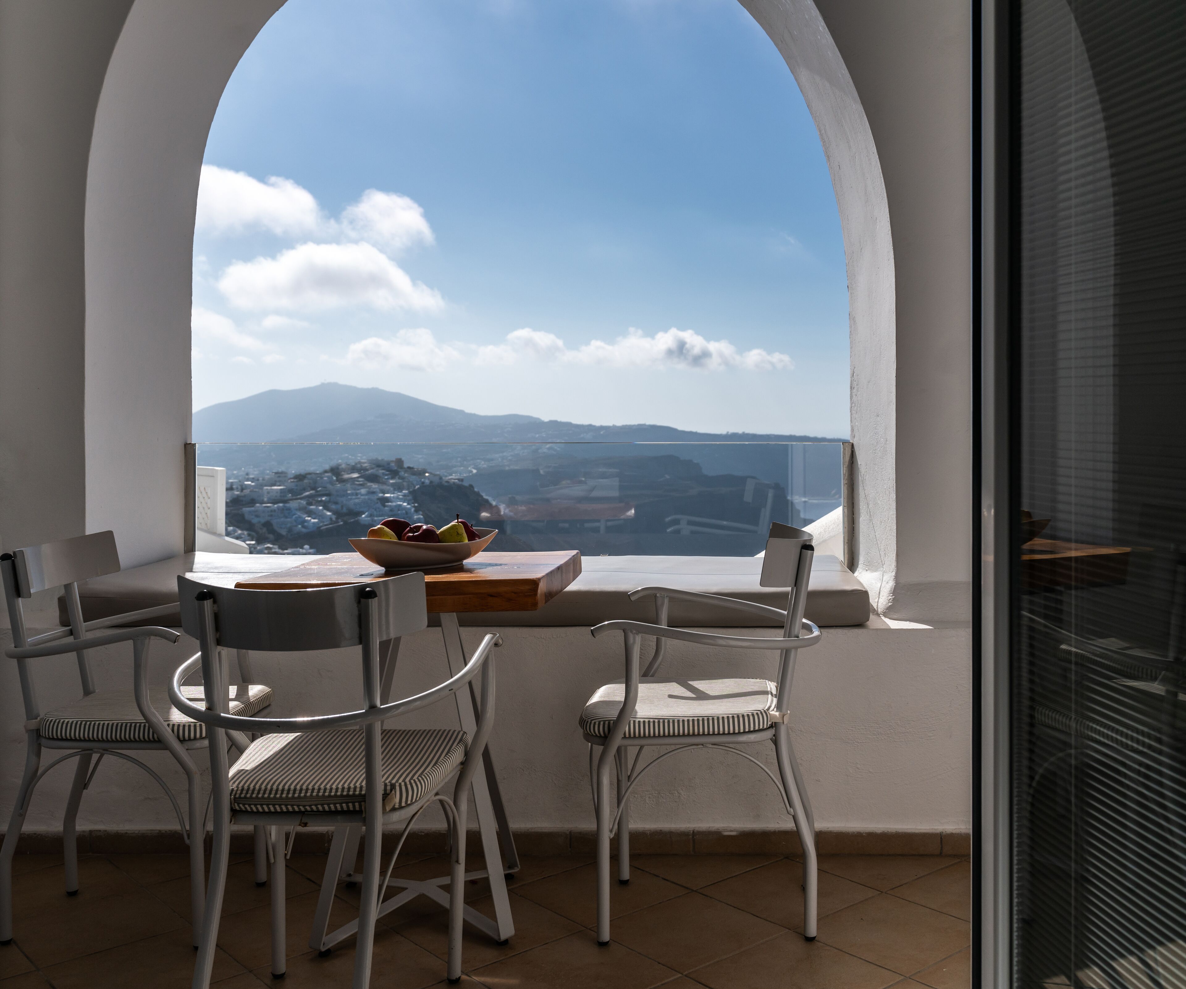 A balcony with a table and chairs, a bowl of fruit, and a view of a coastal town and mountains.