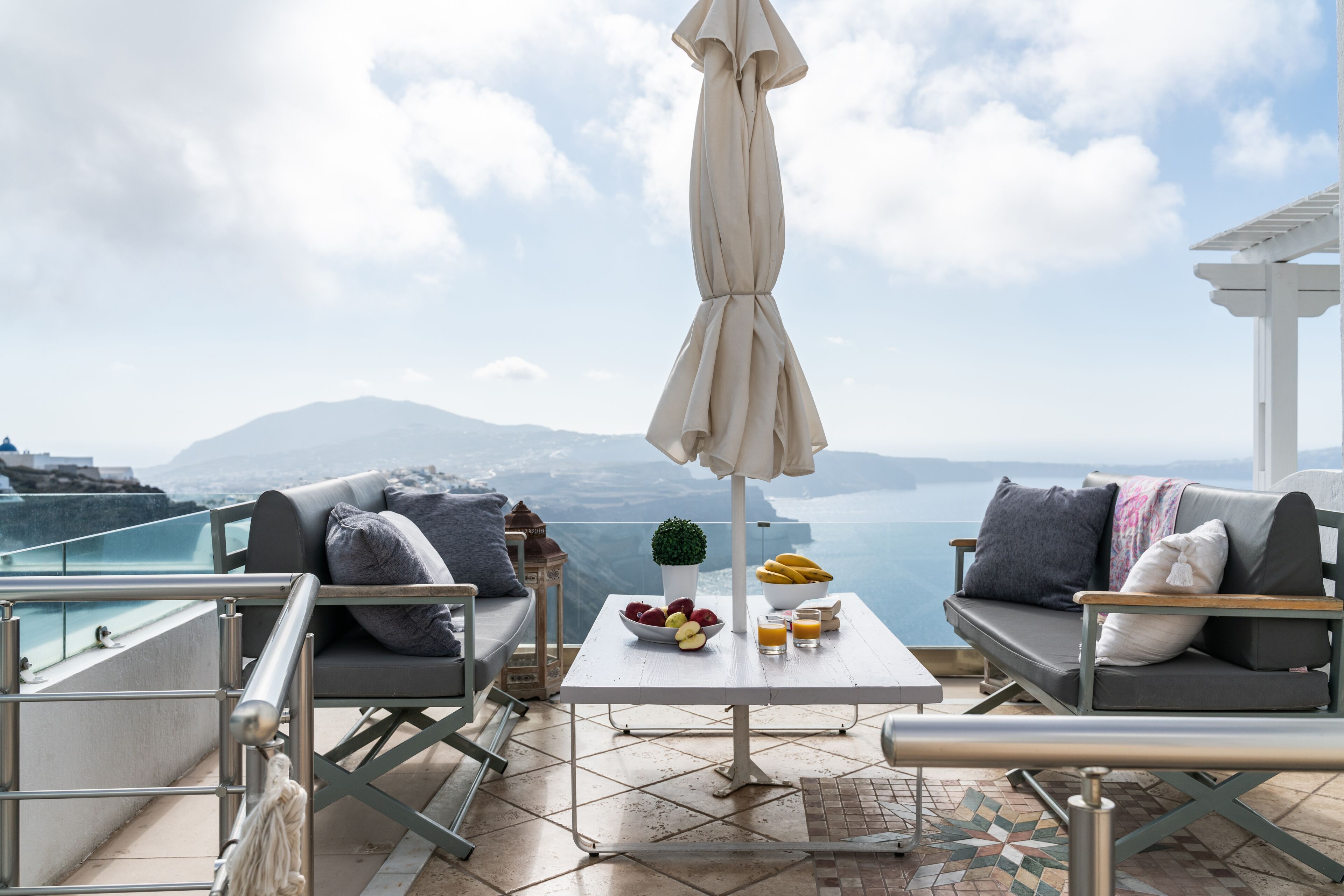 A terrace with a white table, two grey sofas, a potted plant, and a view of the sea and mountains.