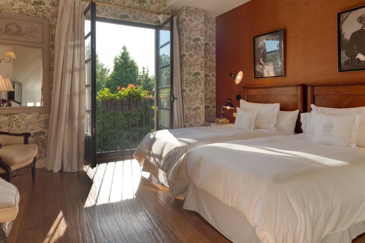 Un dormitorio con dos camas, cabeceros de madera, una mesita de noche con lámpara, una silla y una ventana con cortinas.