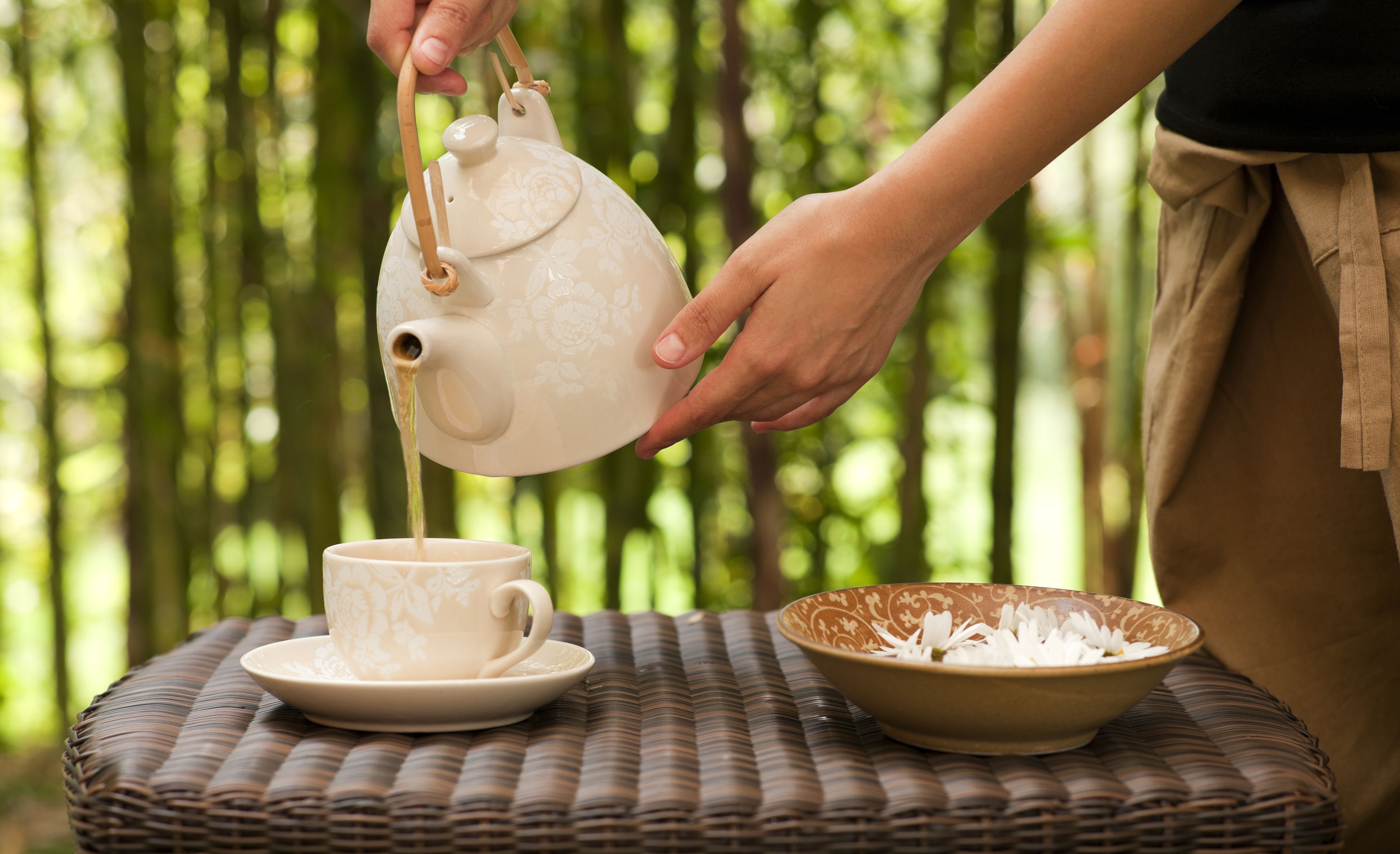 A person pouring tea from a teapot into a cup on a woven table.