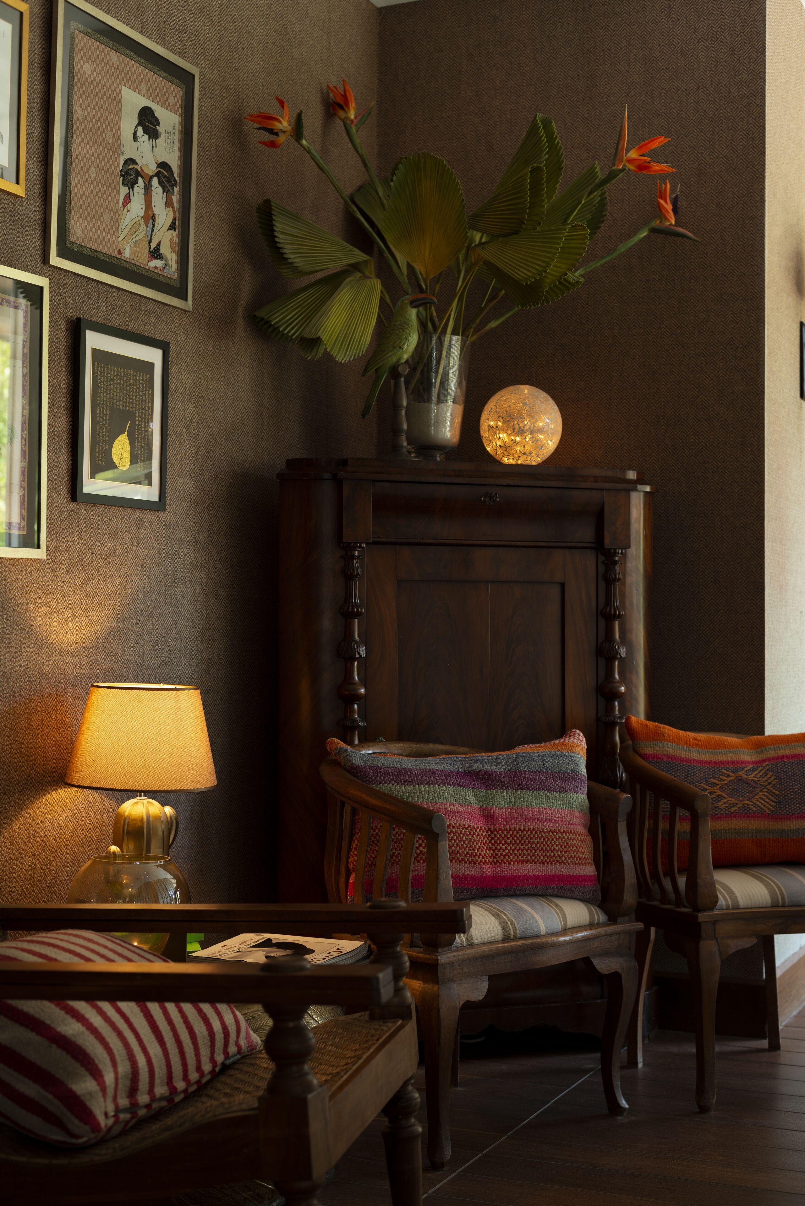 Una habitación acogedora con una cama de madera, una silla, una mesa y una lámpara. Hay cuadros en la pared y una planta en maceta con flores.