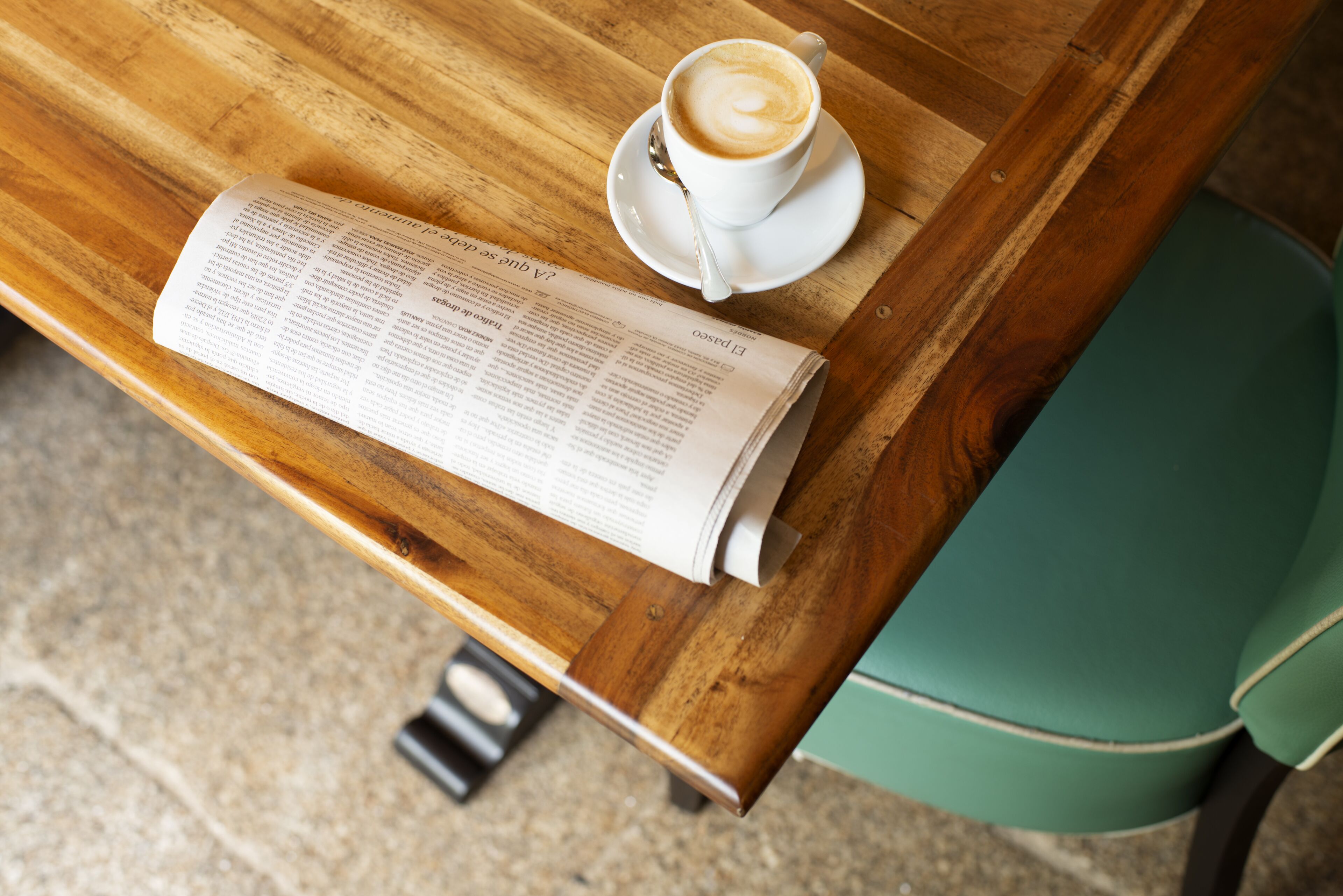 A wooden table with a white cup of coffee, a spoon, and a newspaper.