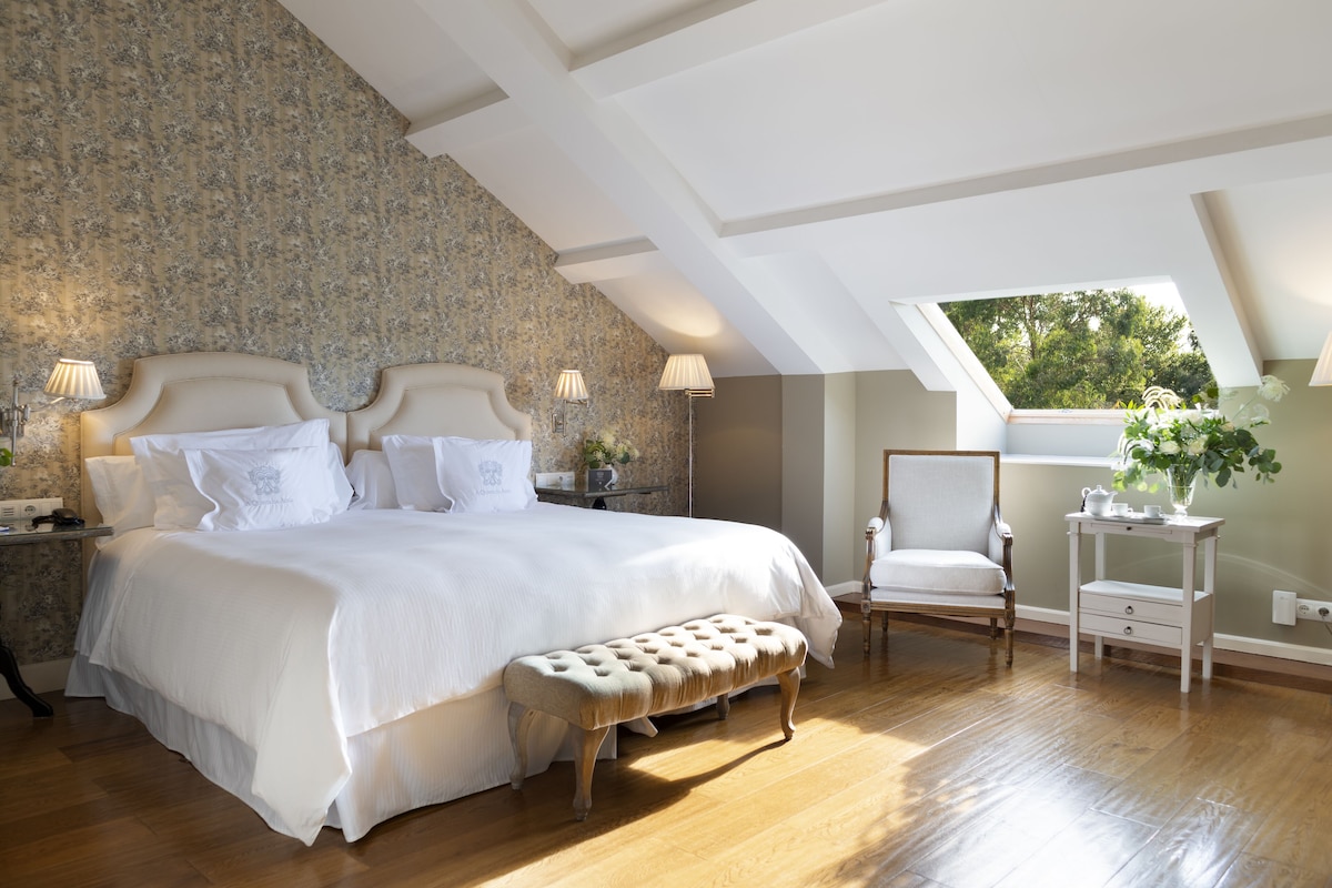 Une chambre à coucher avec un grand lit, un banc, une chaise, une table de chevet et une fenêtre donnant sur des arbres.