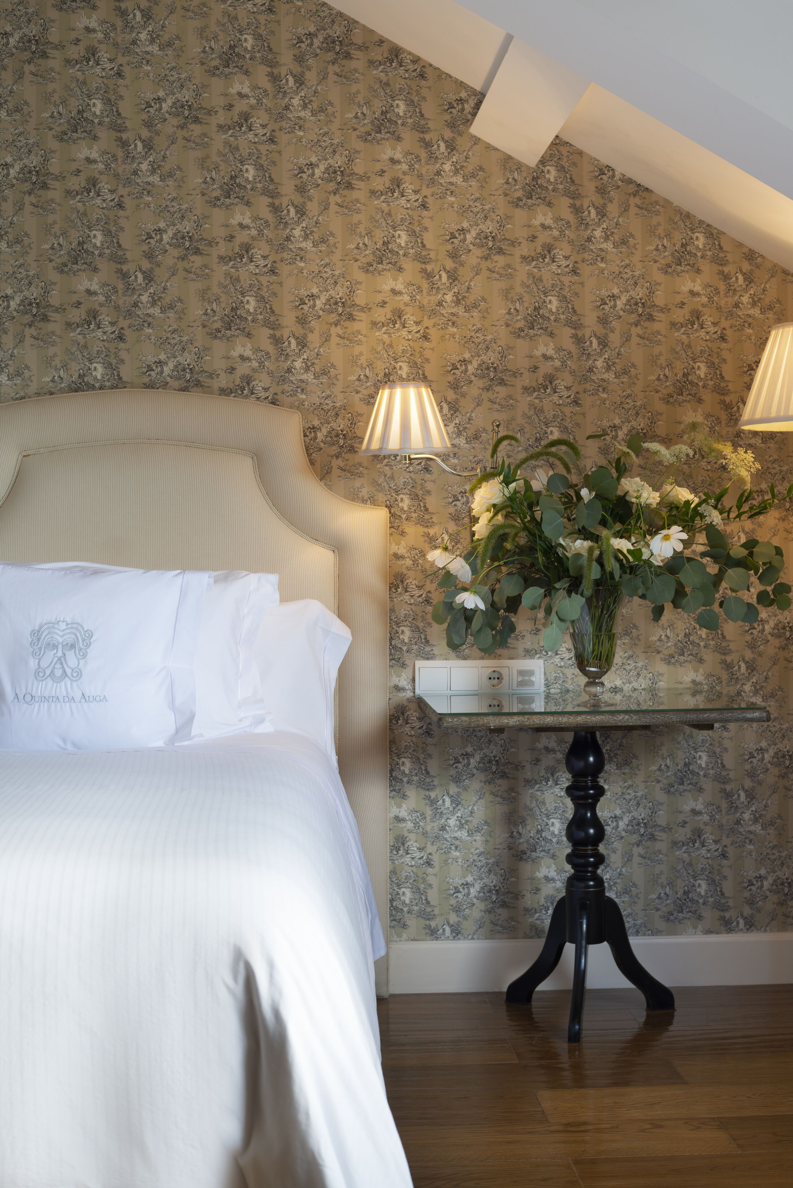 A bedroom with a bed, floral wallpaper, and a bedside table with a vase of flowers.