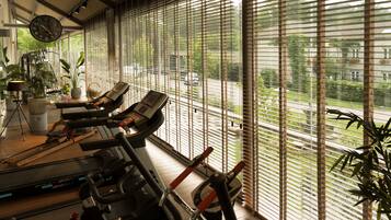 Ein Fitnessraum mit Ergometer und Laufband, großen Fenstern mit Jalousien und einer Wanduhr.