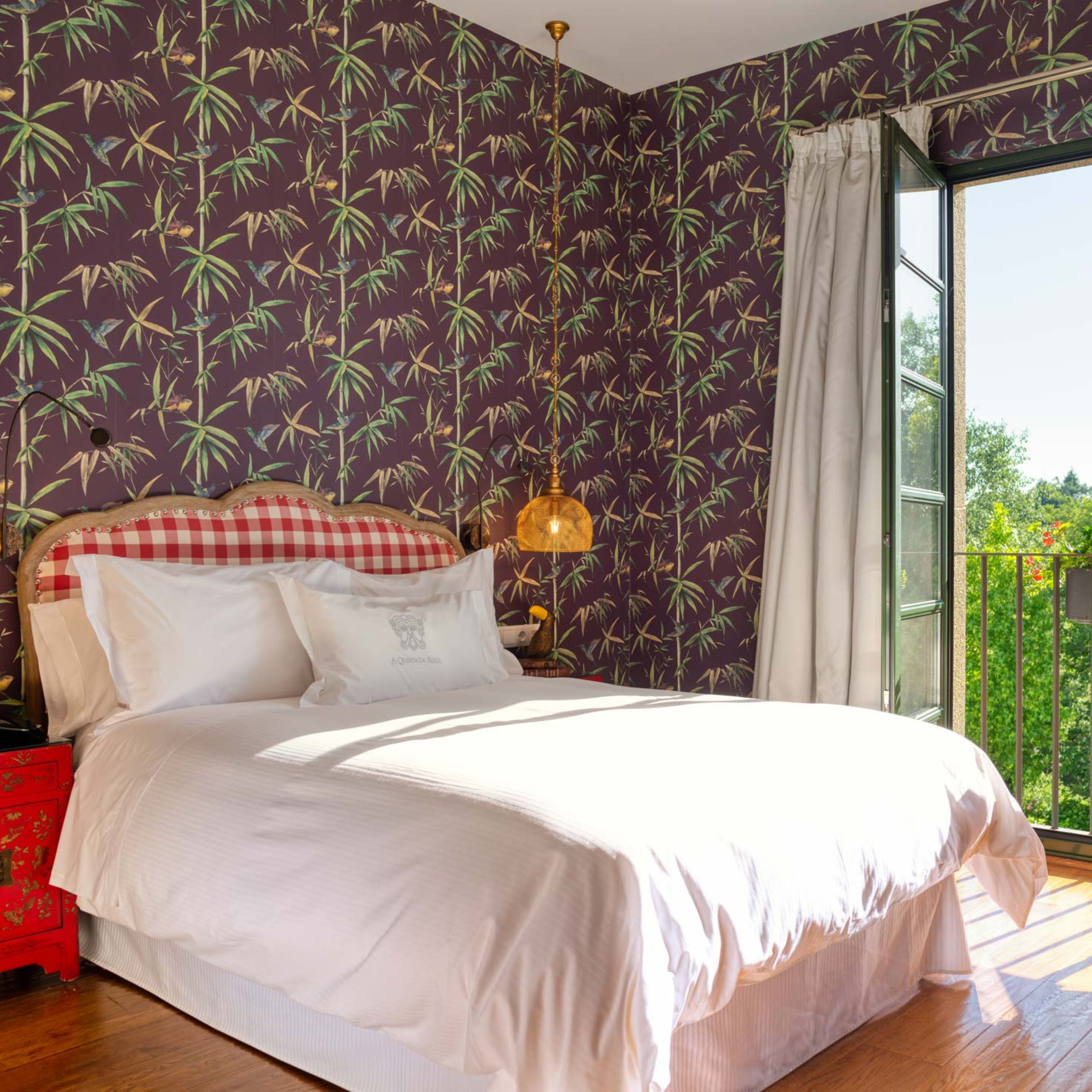 Un dormitorio con una cama, cabecera estampada de cuadros, una lámpara colgante y una ventana con vista a follaje.