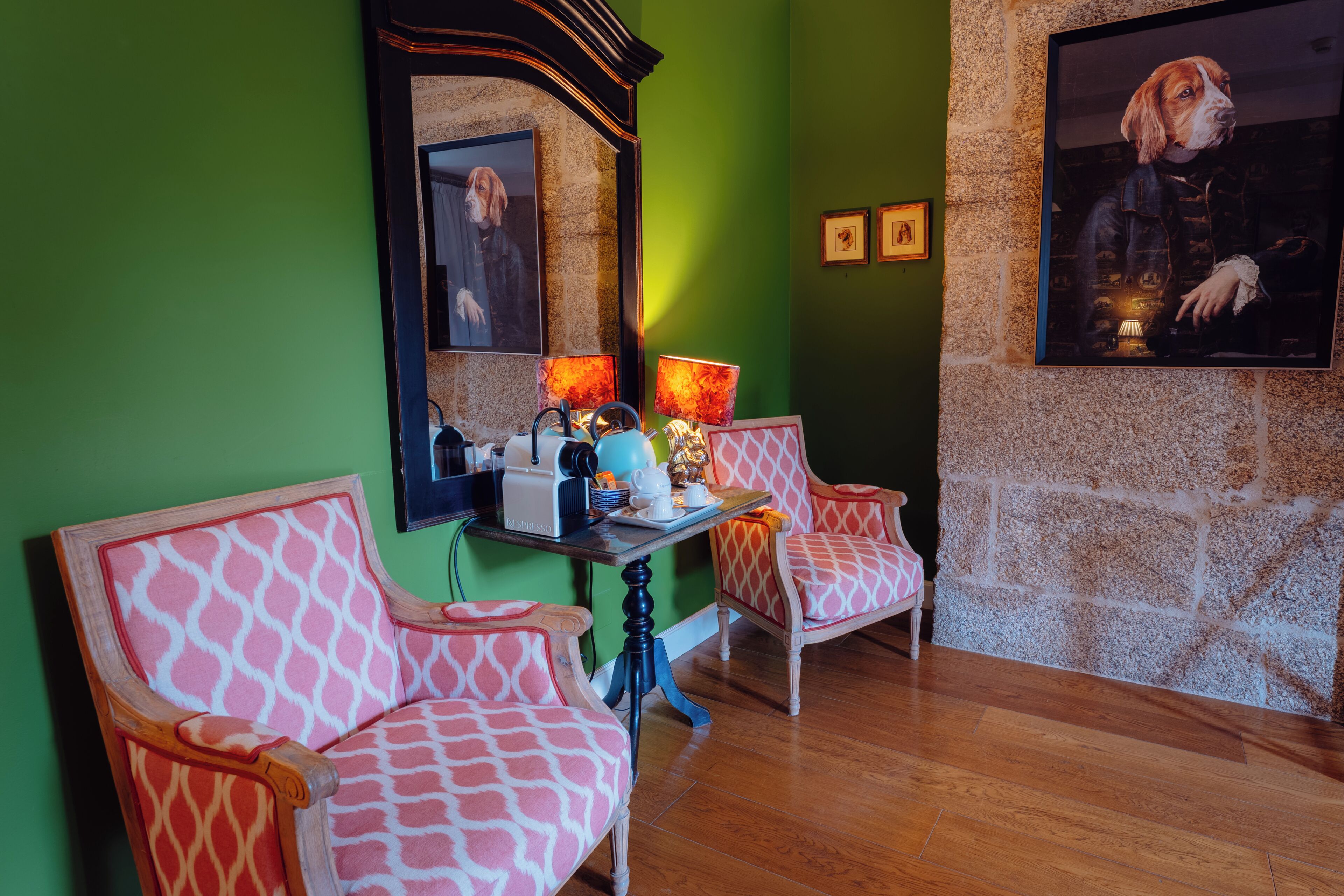 Una habitación con paredes verdes, suelo de madera, dos sillas estampadas, una mesa con una cafetera y un cuadro en la pared.