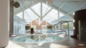 Une grande piscine intérieure dotée d’un haut plafond et de fenêtres de toit, entourée d’un décor intérieur blanc et offrant une vue sur un bâtiment à l’extérieur.