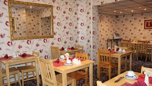 Un coin repas avec des tables et des chaises en bois, un papier peint à motifs floraux et un grand miroir au mur.