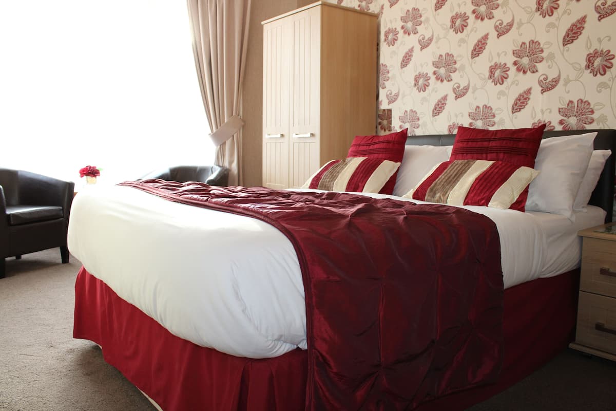 A neatly made bed with a red and white bedspread, a wooden wardrobe, and a floral wallpaper pattern.