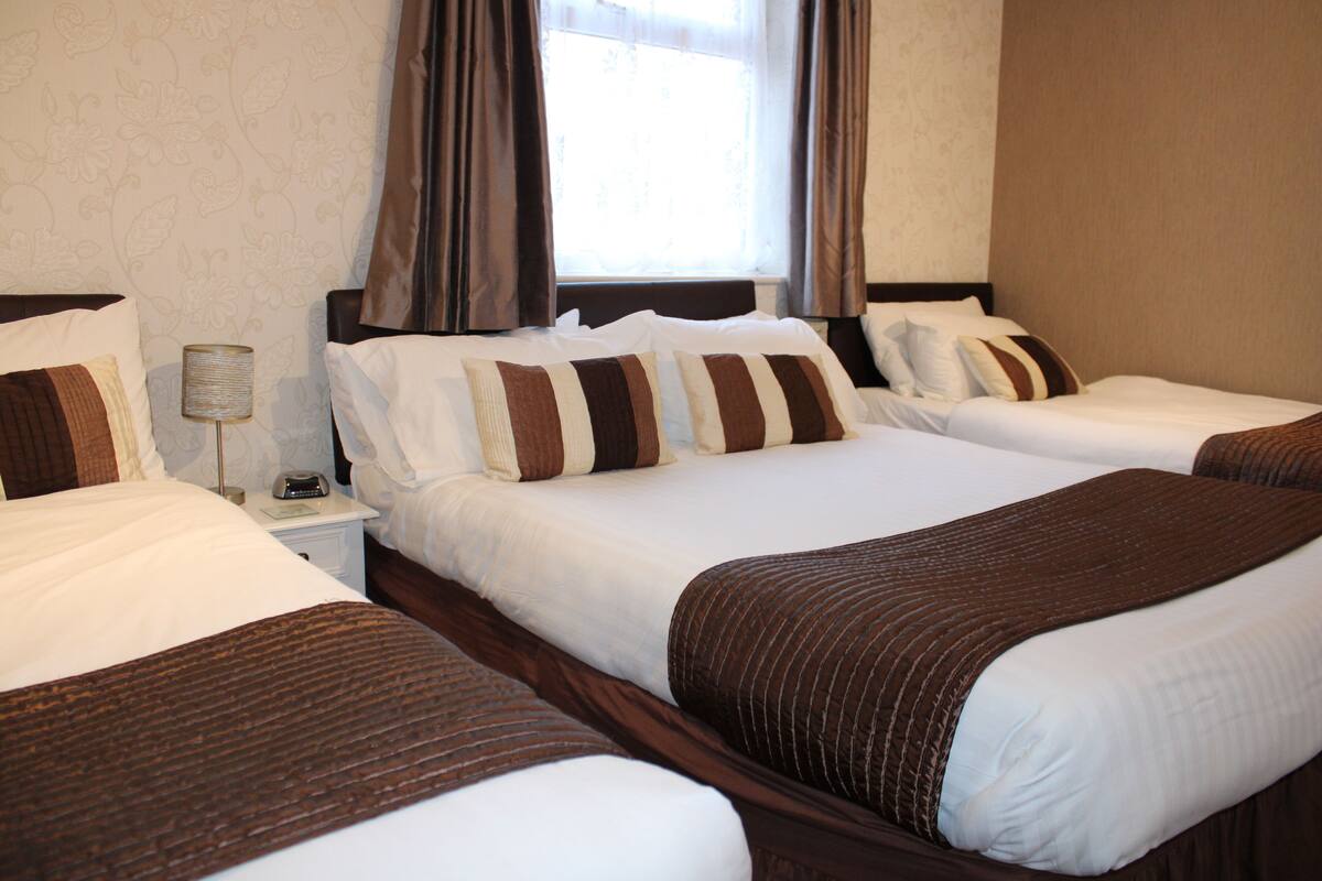 A hotel room with three beds, each with white bedding and brown striped pillows. There is a nightstand with a lamp and a telephone.