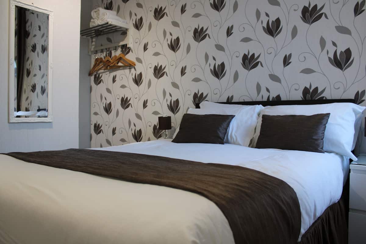 A neatly made bed with a dark brown bedspread and white linens, a wooden hanger rack with clothes, and a mirror on the wall.