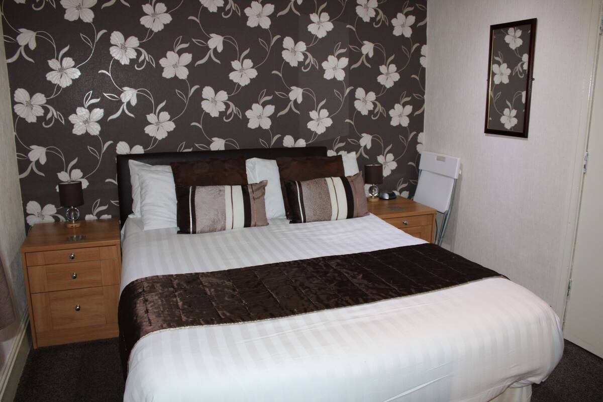 A neatly made bed with a dark brown headboard and white bedding, a wooden nightstand with a lamp, and a floral-patterned wallpaper.