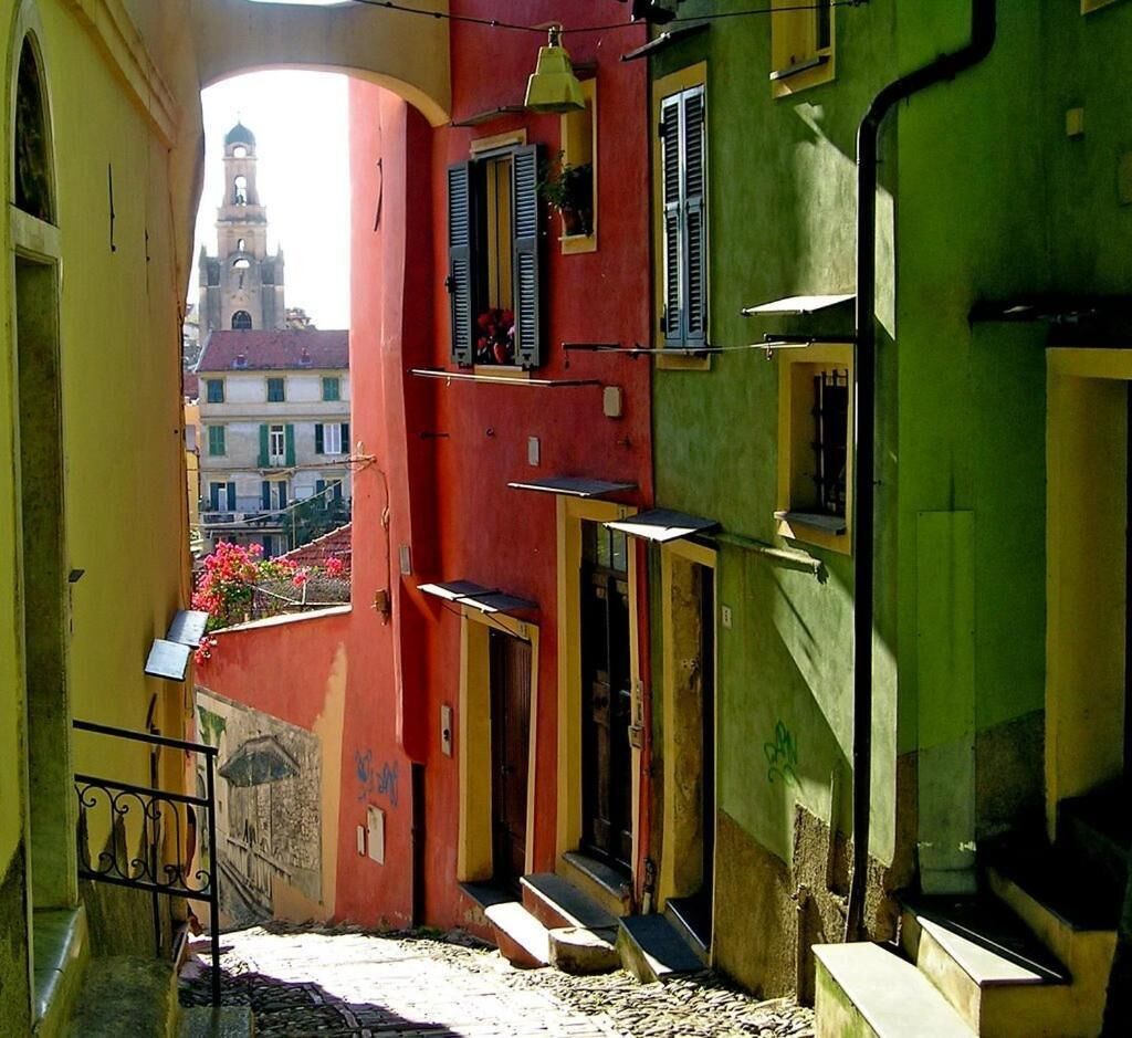 Eine enge Straße mit bunten Häusern, einem sichtbaren Glockenturm und Stufen, die zu einem Hauseingang führen.