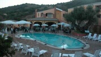 Une piscine entourée de bâtiments et de collines, avec des chaises longues et des parasols.