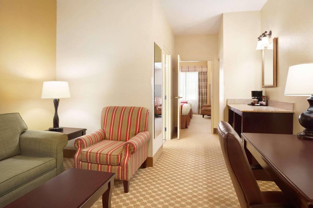 Une chambre d’hôtel avec un tapis à carreaux, un canapé vert, un fauteuil à rayures, une table en bois, un bureau et un miroir.