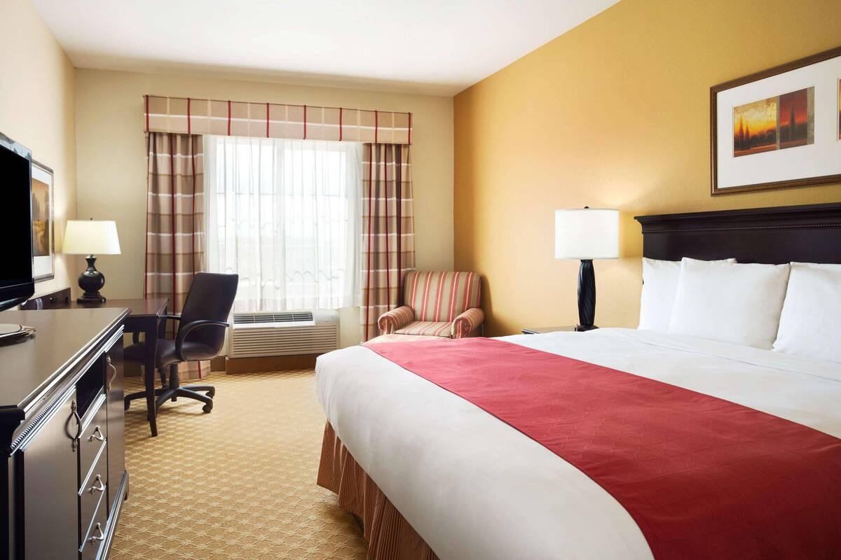 Une chambre d’hôtel avec un grand lit, un bureau avec une lampe, une chaise et une fenêtre avec des rideaux.