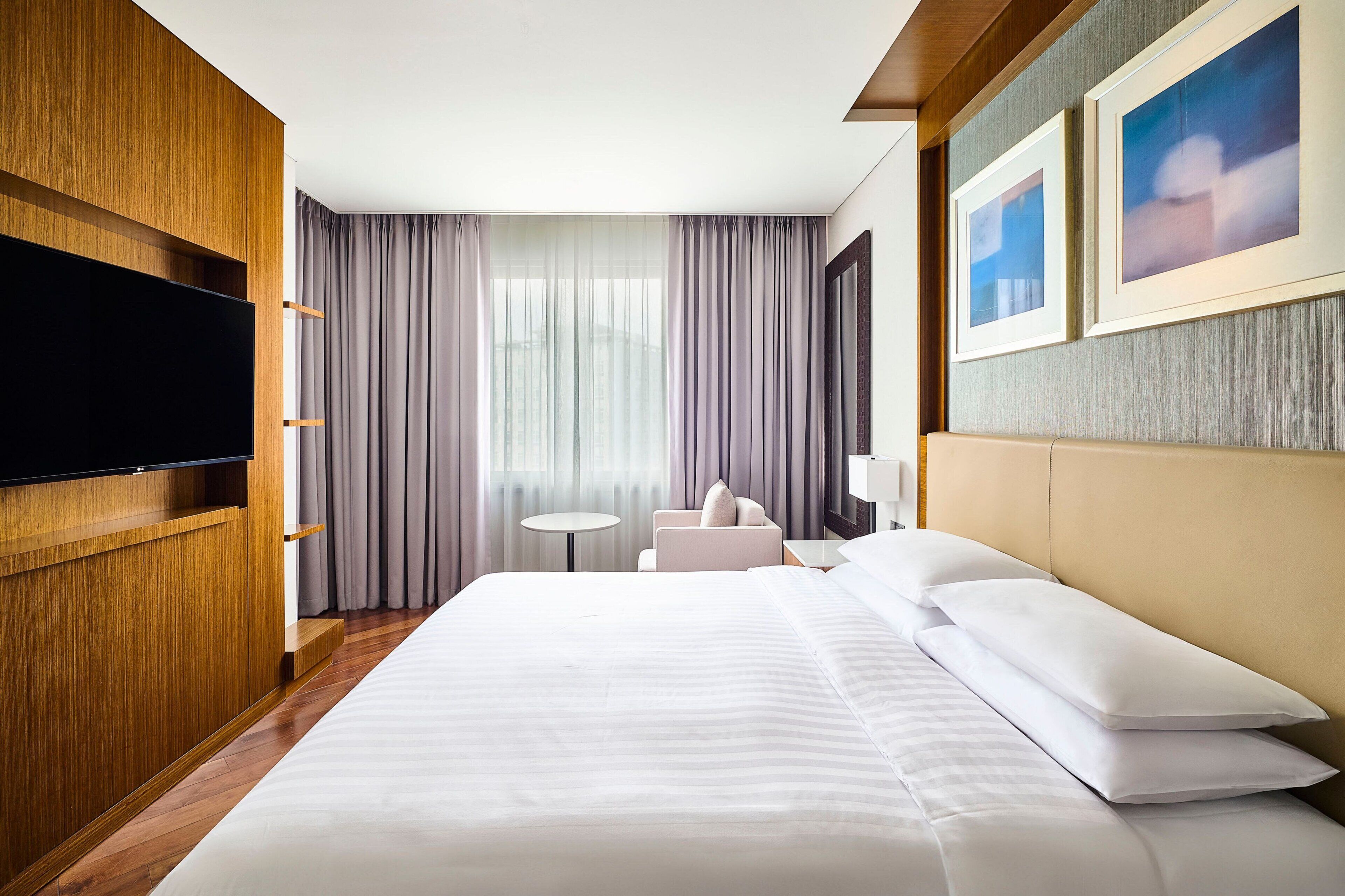 A hotel room with a large bed, a flat-screen TV mounted on a wooden panel, a small round table, a sofa, and a large window with curtains.