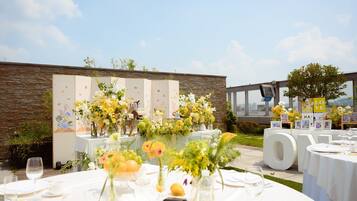 An outdoor wedding reception with tables set for guests, floral centerpieces, and a backdrop with a stone wall and framed pictures.