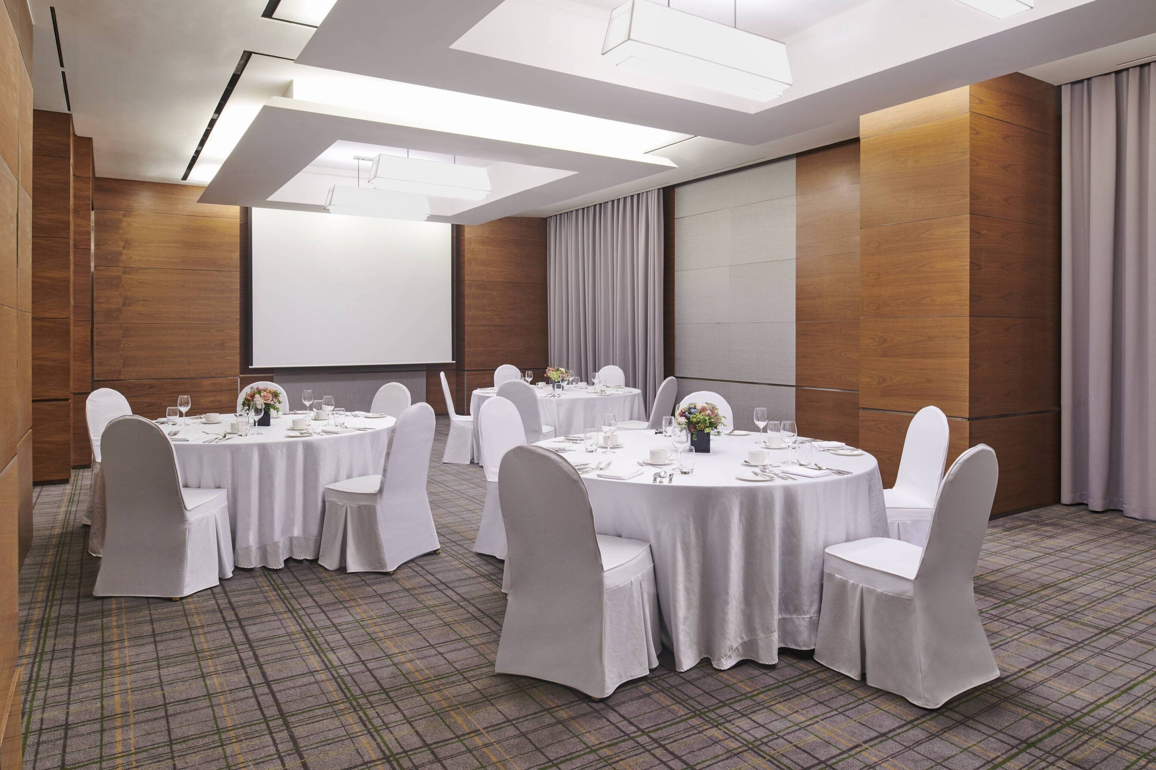 Un salón de conferencias con mesas redondas, sillas blancas y una pantalla grande.