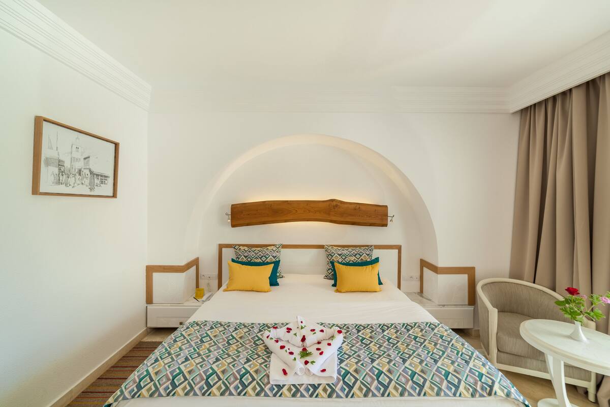 A hotel room with a large bed, two pillows, a wooden headboard, a small table with a vase, and a chair.