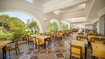 A spacious dining area with multiple tables and chairs, large windows, and a view of greenery outside.