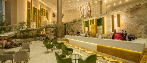 A lobby with a reception desk, green chairs, a marble floor, and a staircase.