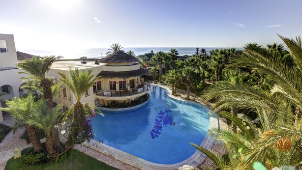 Una piscina en un entorno tropical, rodeada de palmeras y un edificio con estructura circular.