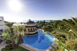 A swimming pool with a tropical setting, surrounded by palm trees and a building with a circular structure. - Magic Life Africana (Hammamet)