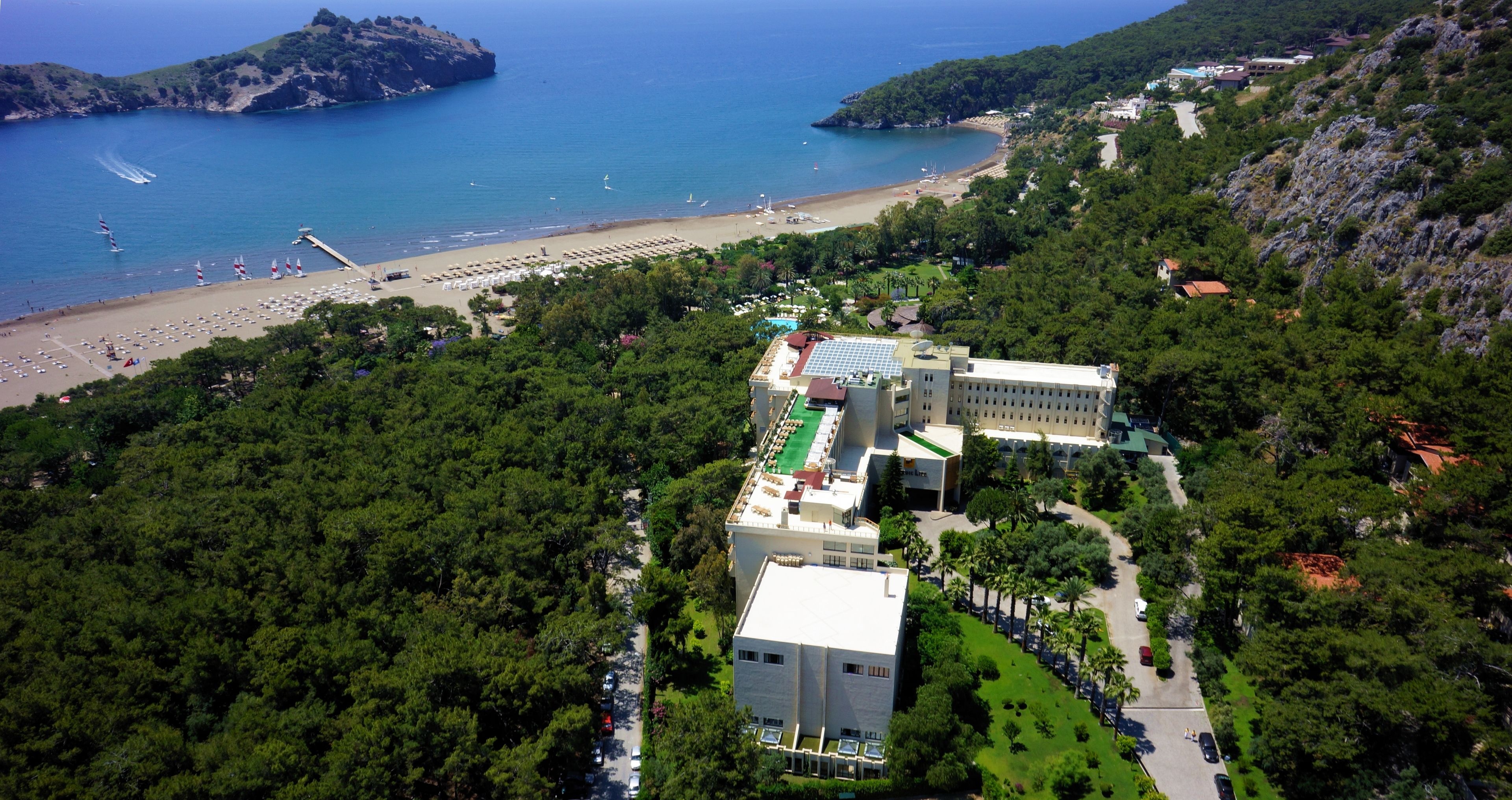 An aerial view of a coastal resort with a beach, dense greenery, and a large building with multiple floors and a rooftop terrace.