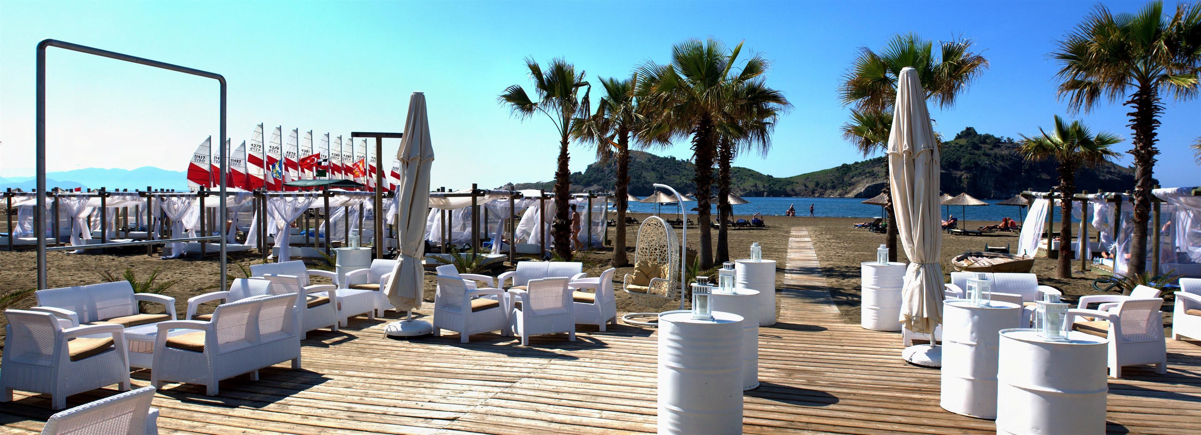 A beachside venue with white wicker chairs and tables, a wooden deck, and a backdrop of flags and palm trees.