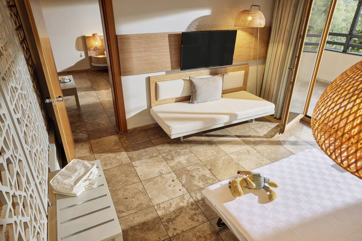 Ein modernes Hotelzimmer mit Bett, Wand-Fernseher, Nachttisch und Blick ins Freie.