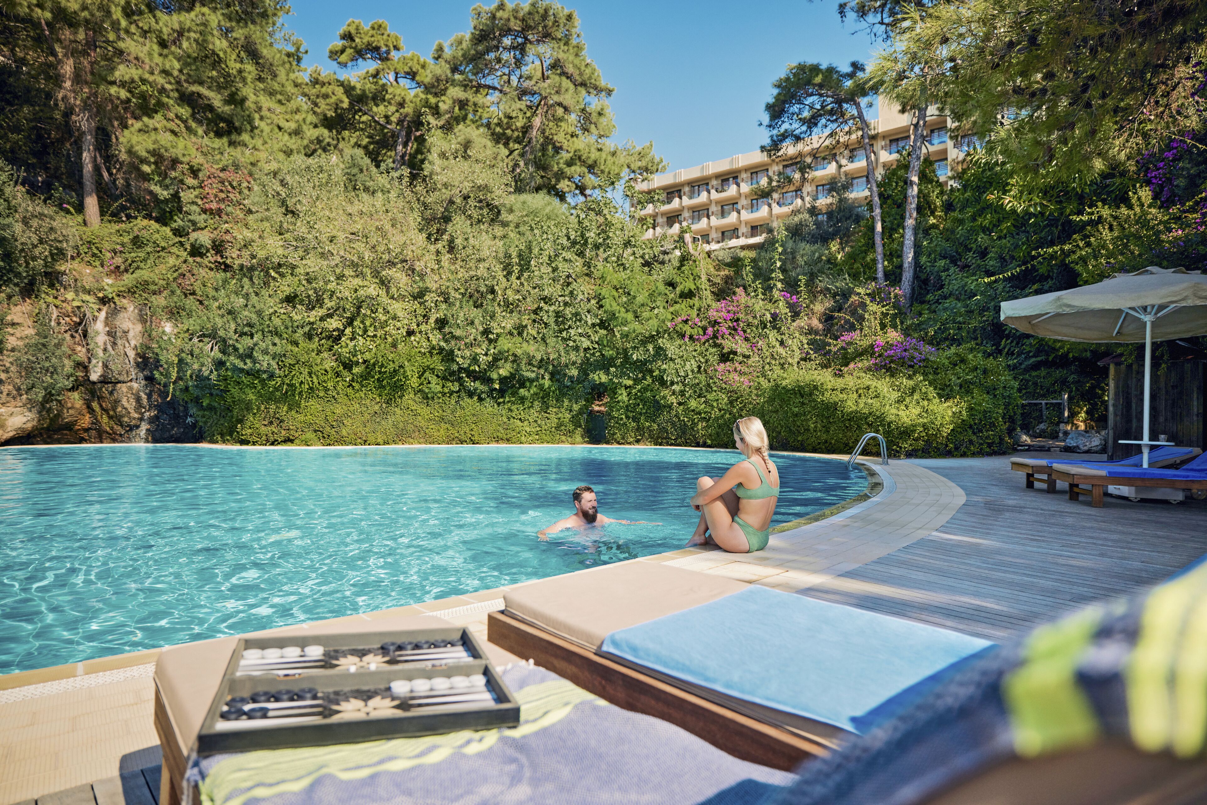 A swimming pool with lounge chairs and a wooden deck, surrounded by lush greenery and a building in the background.