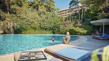 A swimming pool with lounge chairs and a wooden deck, surrounded by lush greenery and a building in the background.