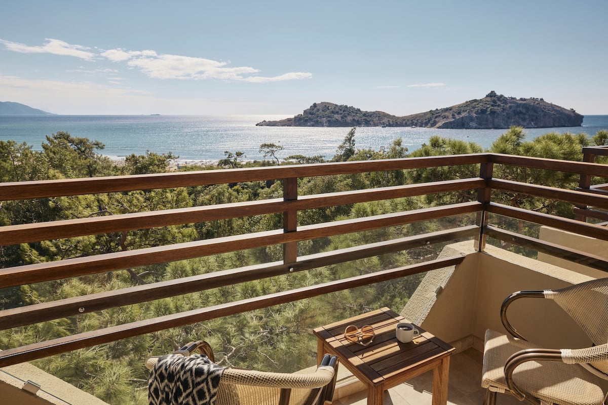 Ein Balkon mit Blick auf das Meer und eine kleine Insel. Es stehen Korbmöbel und ein kleiner Tisch mit einer Sonnenbrille und einer Tasse bereit.