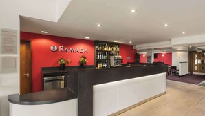 Uma área de recepção com balcão, uma parede vermelha com o logotipo 'RAMADA', um espaço de bar com vários frascos e uma máquina de café.
