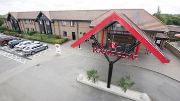 L’entrée d’un hôtel, reconnaissable à une enseigne rouge bien visible, un parking avec plusieurs voitures et la façade d’un immeuble en briques.