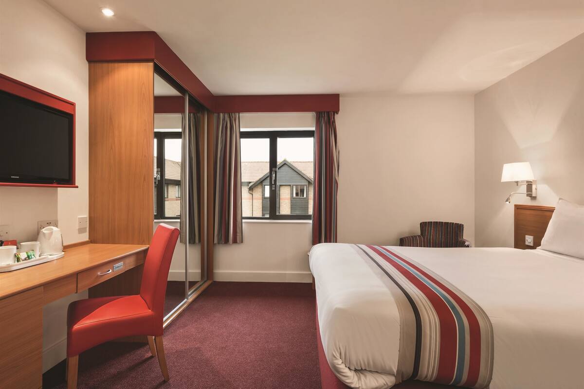A hotel room with a bed, a desk, a chair, a TV, and a window with curtains.