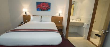 Quarto de hotel com uma cama grande, abajures, um quadro na parede e um banheiro visível por uma porta aberta.