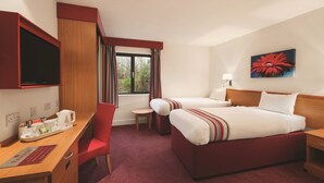 Quarto de hotel com uma mesa, duas camas, televisão, janela com cortinas e um quadro na parede.