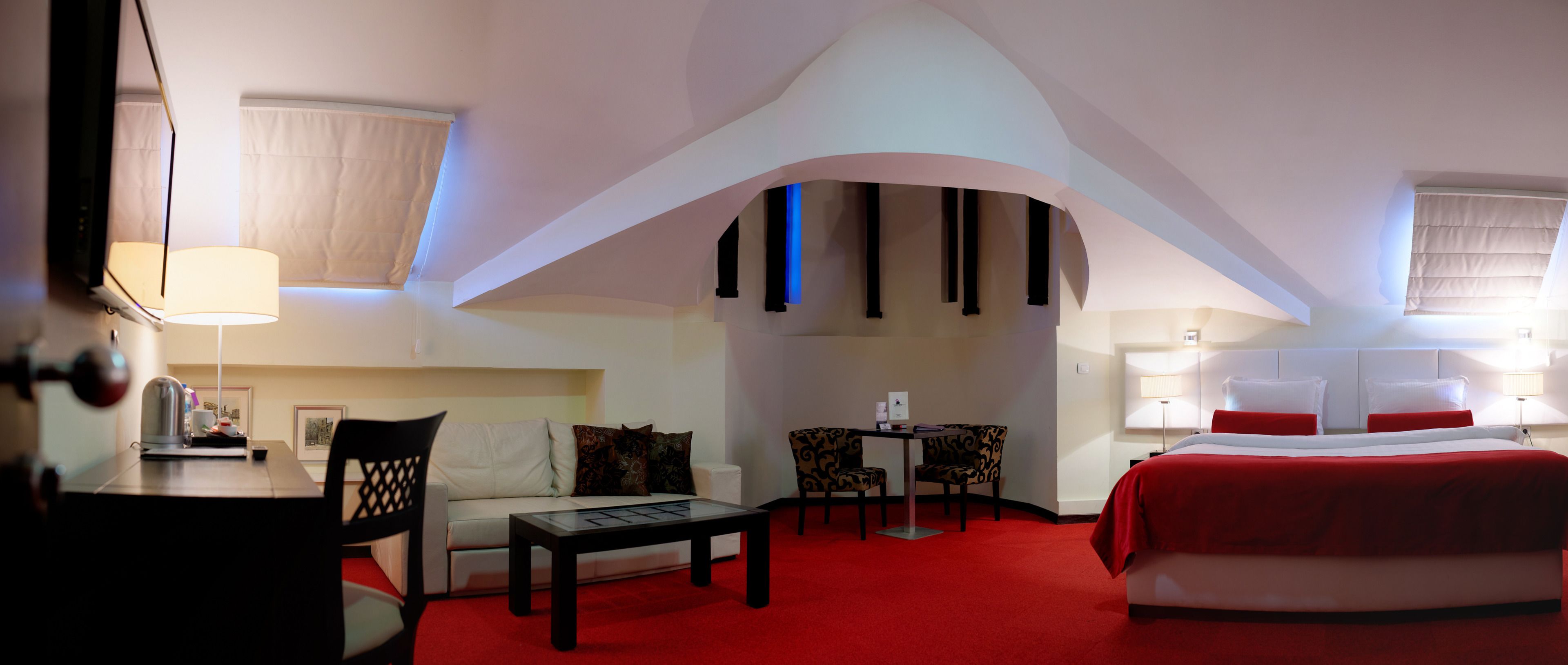 A hotel room with a red carpet, a bed with red bedding, a sofa, a table with chairs, and a TV.