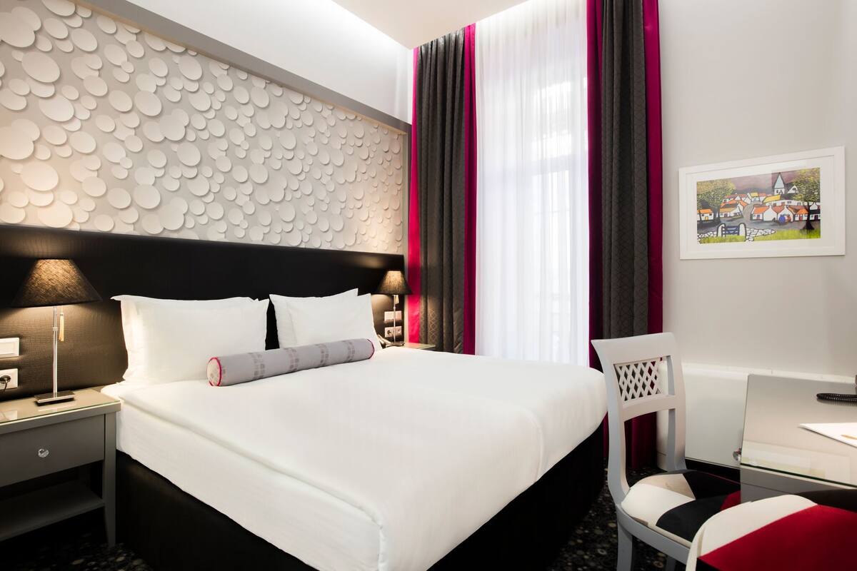 Una habitación de hotel con una cama, una mesita de noche con lámpara, una silla y un escritorio con computadora.