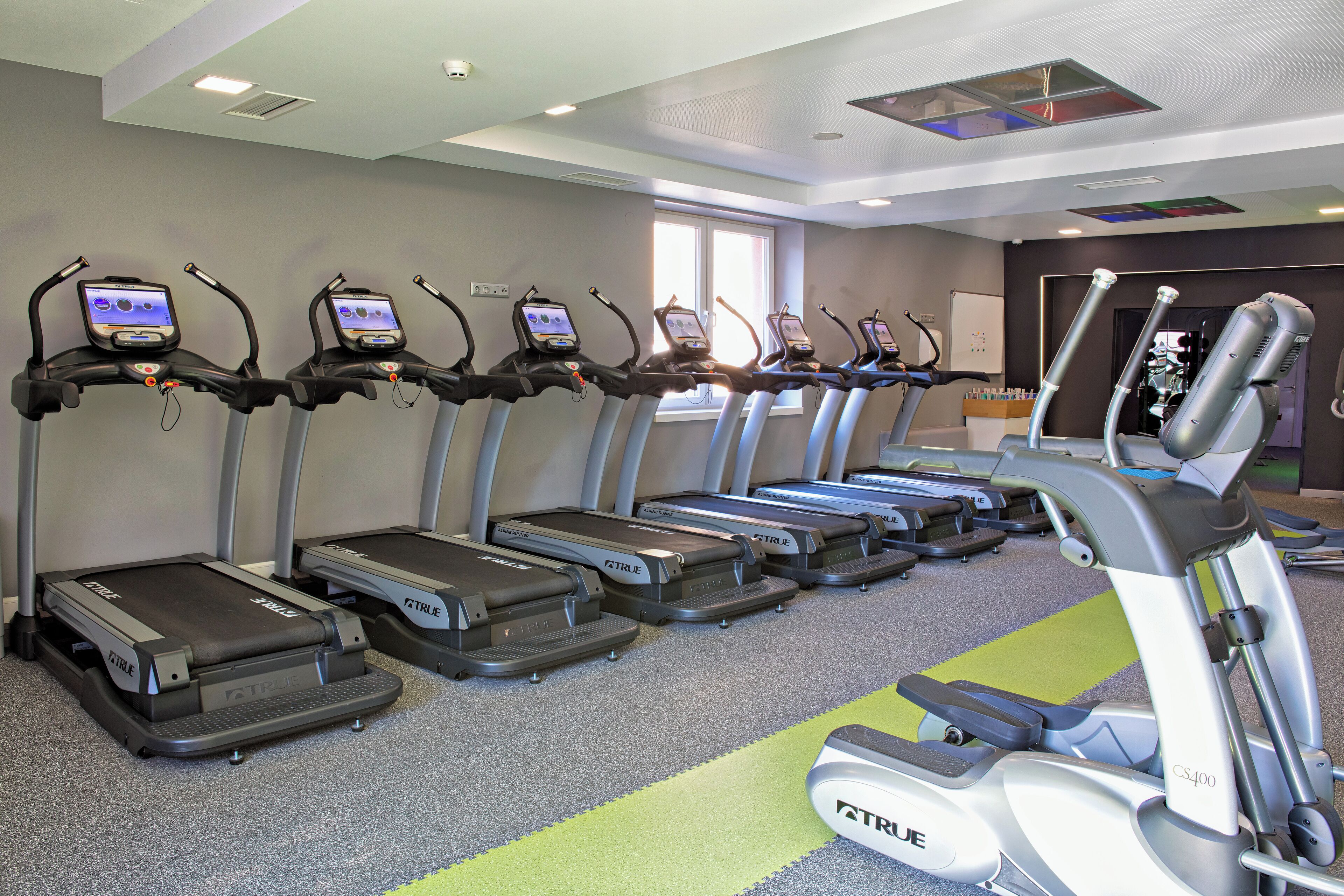 En rad löpband med digitala displayer, placerade i ett gym med ett grått golv och ett fönster i bakgrunden.