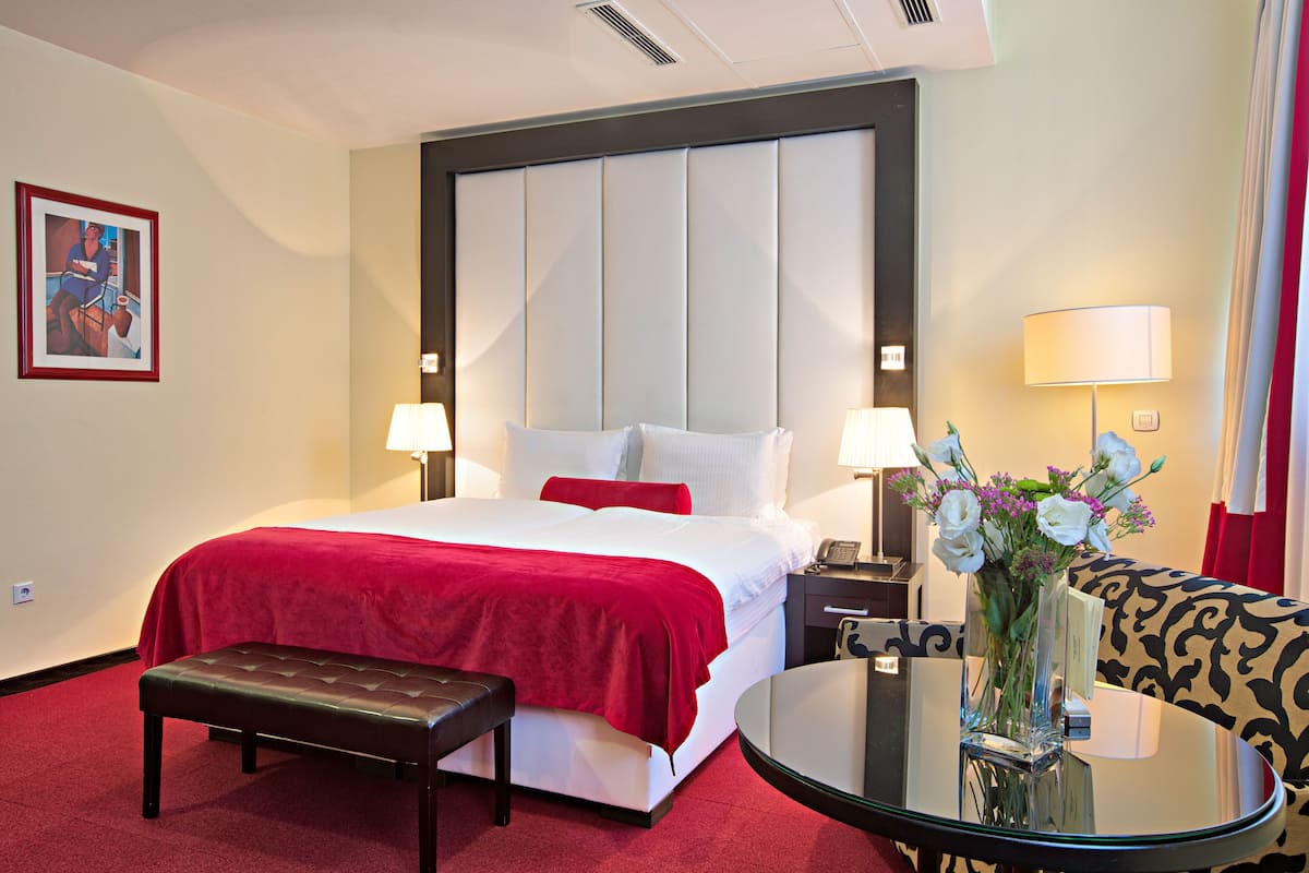 Un cuarto de hotel con una cama grande, dos mesitas de noche con lámparas, una pequeña mesa con un florero, un banco y un sofá estampado.