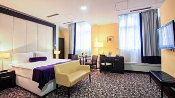 A hotel room with a bed, a desk, a chair, a sofa, a TV, and a window with curtains.