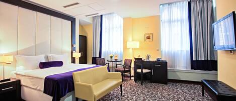 A hotel room with a bed, a desk, a chair, a sofa, a TV, and a window with curtains.