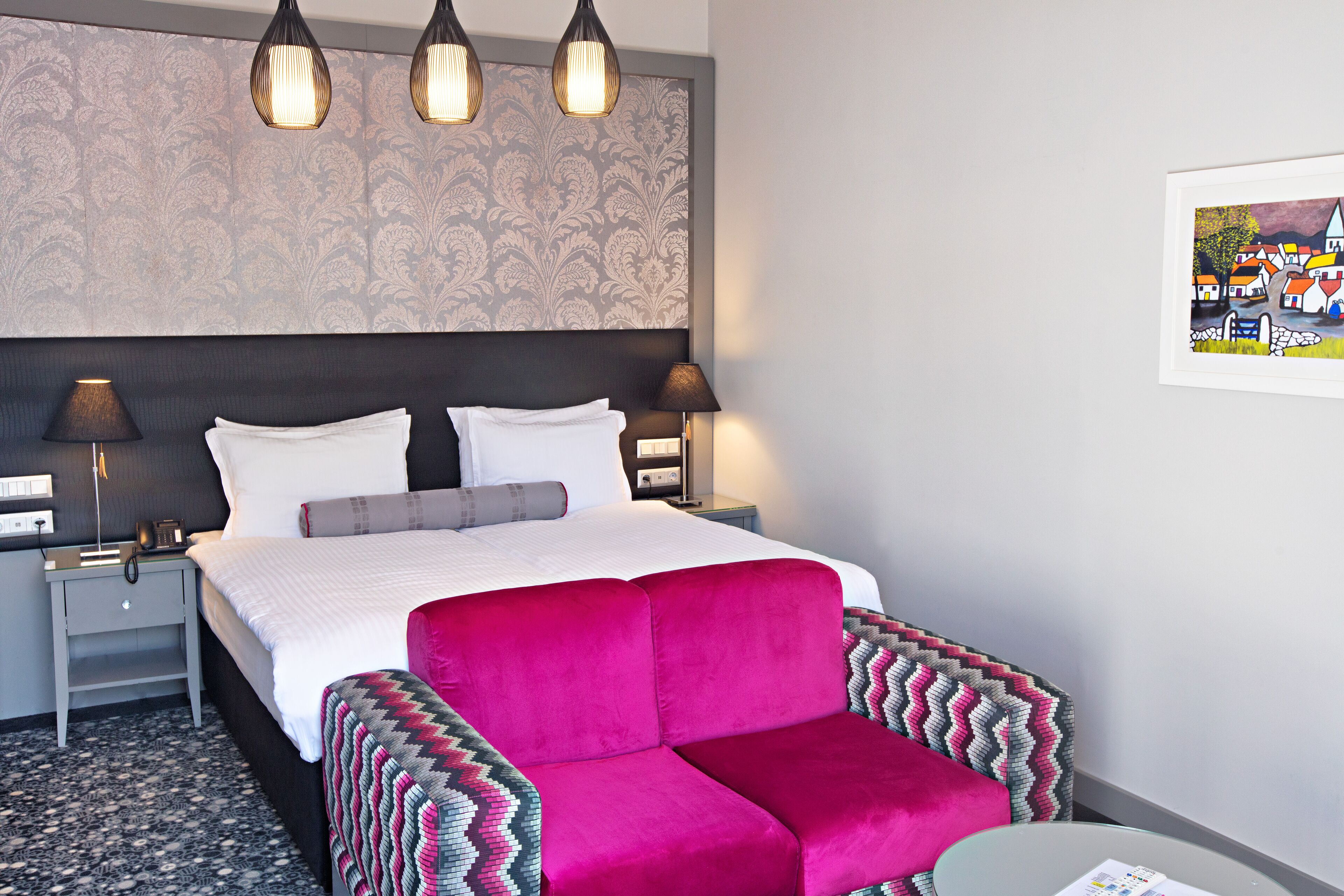 A hotel room with a large bed, two bedside lamps, a patterned wallpaper, and a framed artwork on the wall.