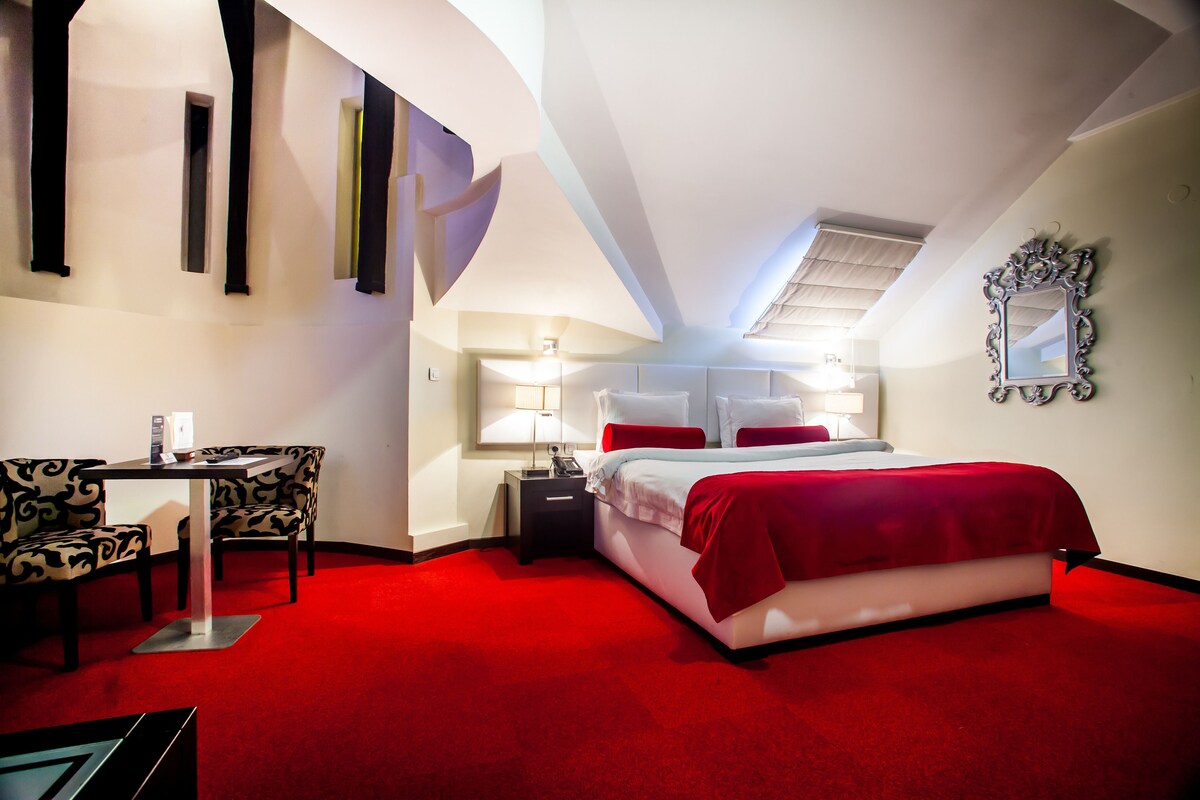 A hotel room with a red carpet, a bed with white and red bedding, a desk with a chair, and a mirror on the wall.