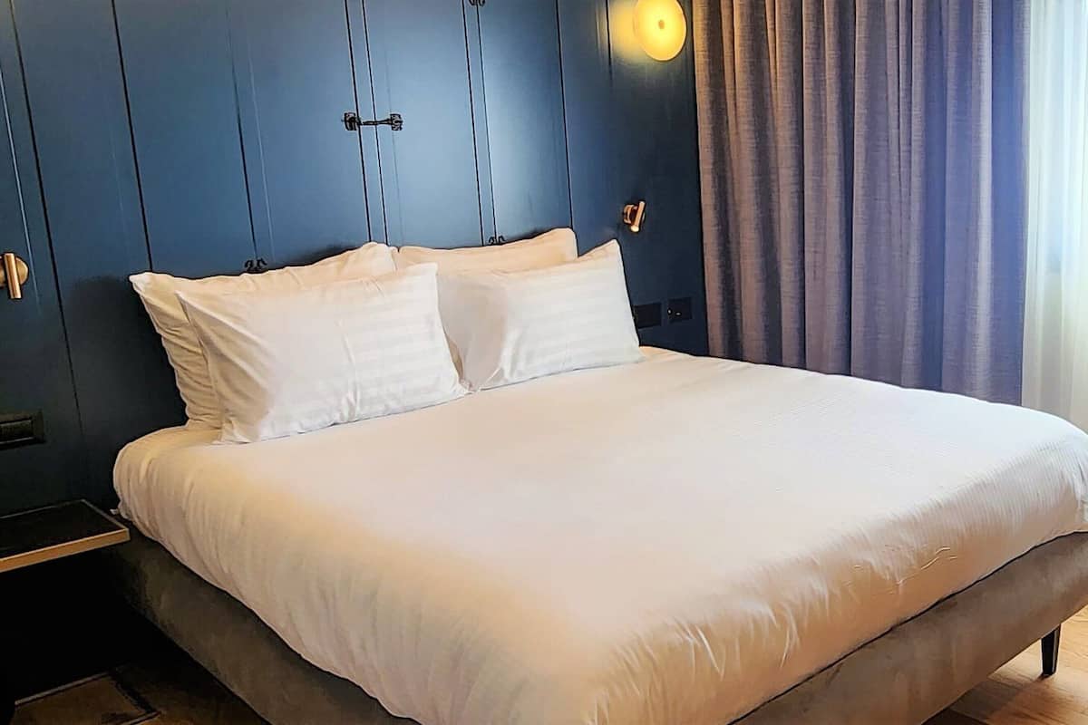 A neatly made bed in a room with a dark blue wall, a small bedside table, and a chair.